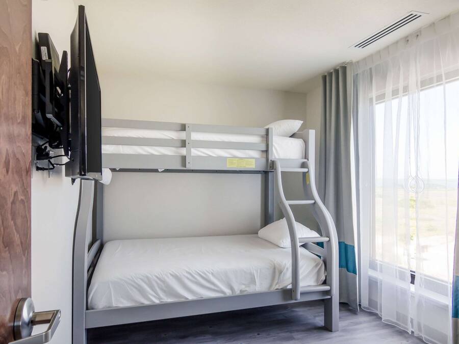A bunk bed room with a TV mounted on the wall, a window with sheer curtains, and a wooden door.