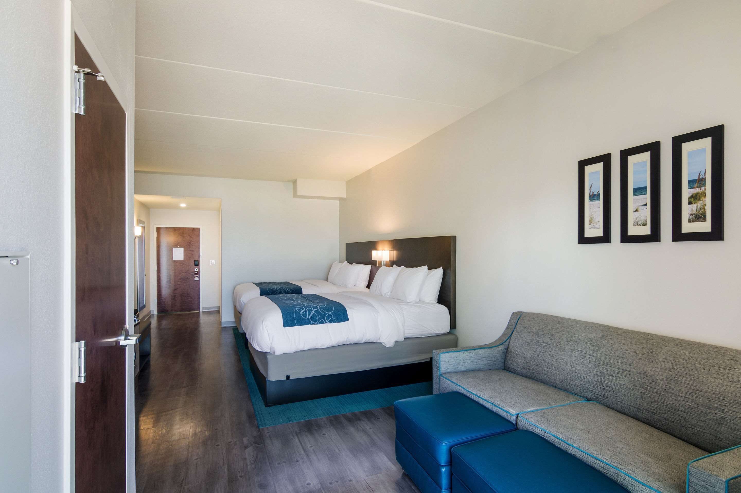 A hotel room with a bed, a sofa, and a small table. There is a door on the left and a hallway leading to another room.