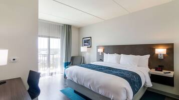 A hotel room with a large bed, a desk with a chair, a bedside table with a lamp, and a window with curtains.