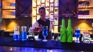 Eine Bar mit einem Barkeeper, blauen und grünen Flaschen sowie Gläsern auf einer Barauflage.