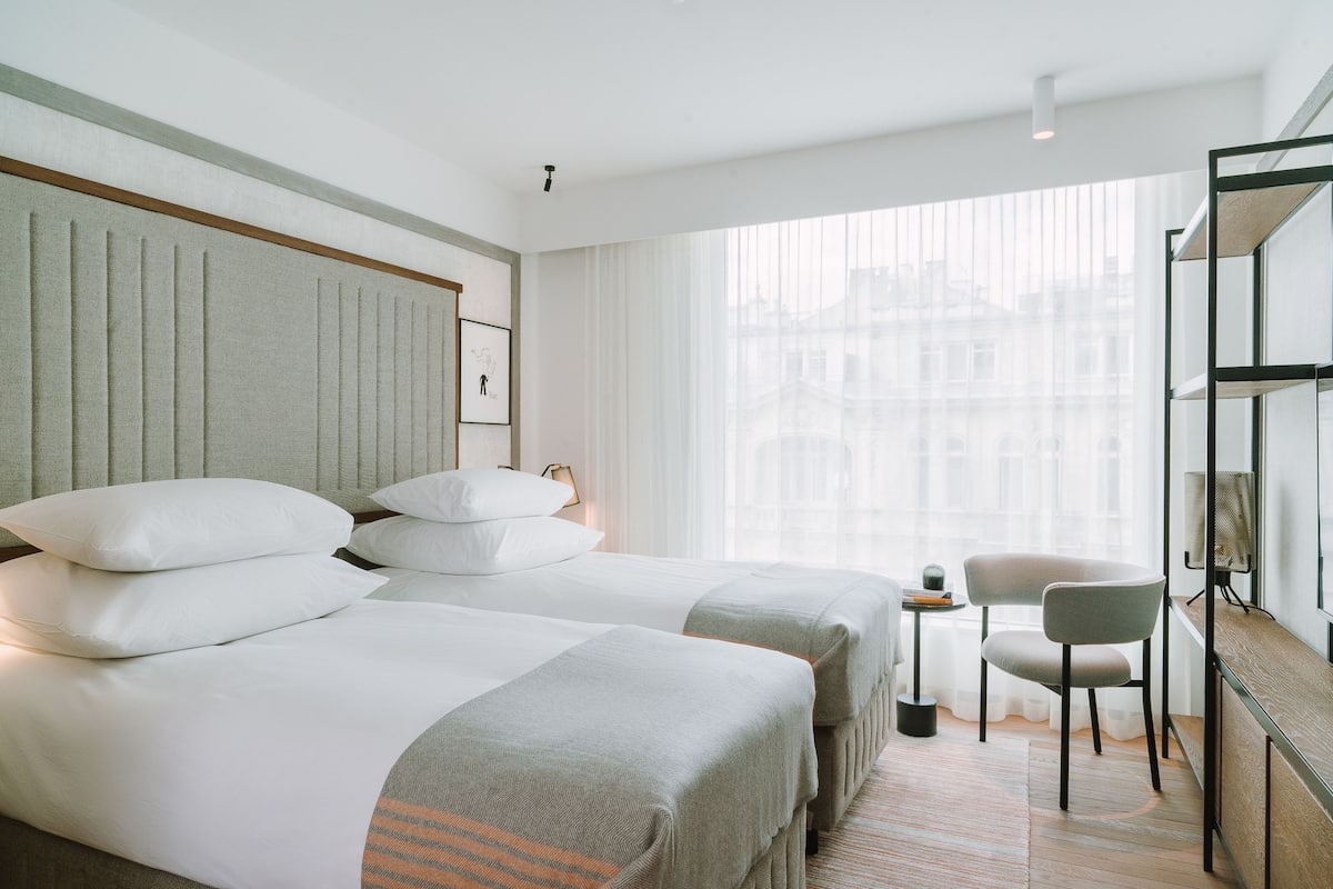 Ein Hotelzimmer mit zwei Betten, einem kleinen Tisch, einem Sessel und einem großen Fenster mit Blick auf die Stadt.