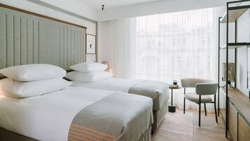 A hotel room with two beds, a small table, a chair, and a large window with a city view.
