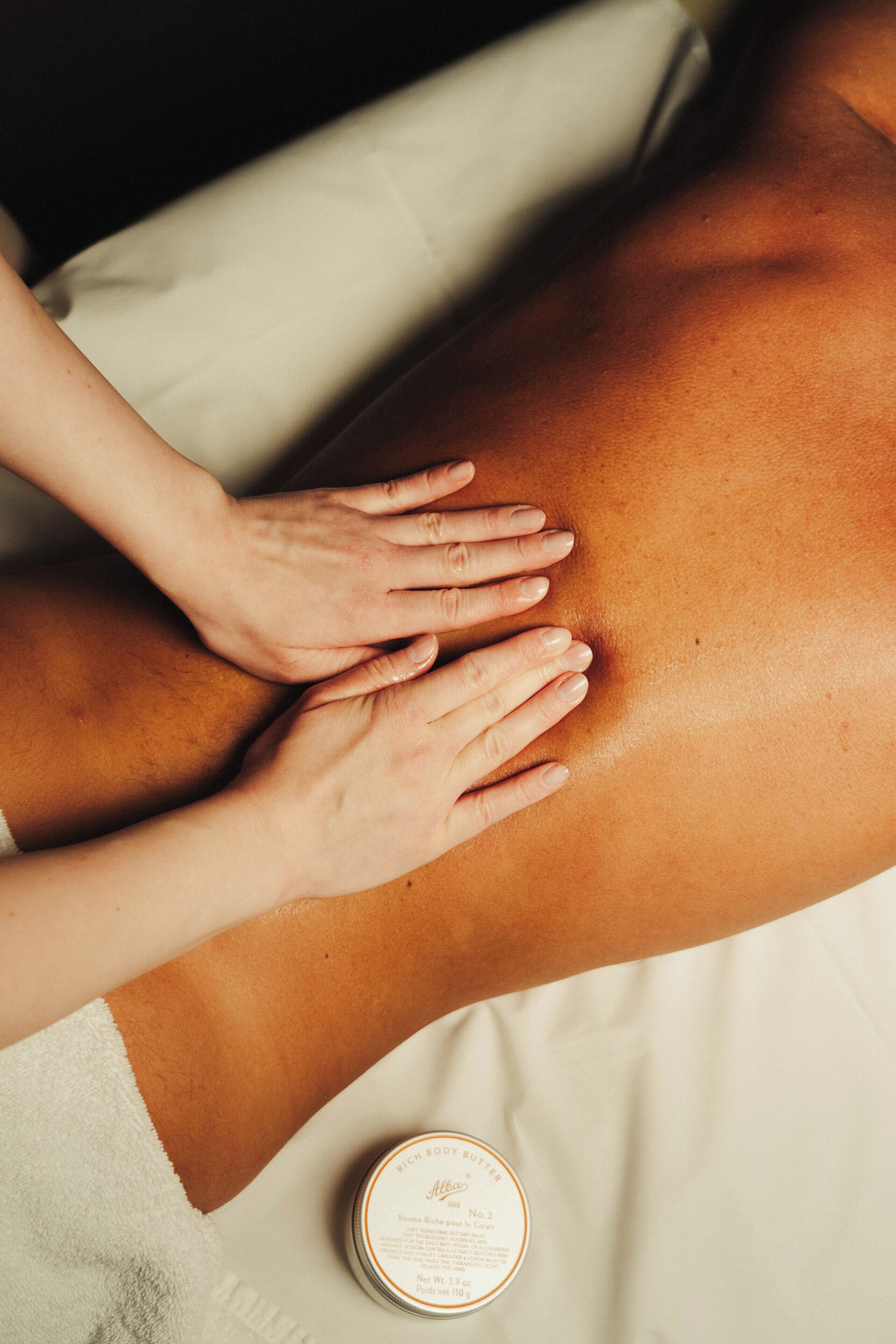 A close-up of a person's back being massaged with hands, a white towel, and a small container of lotion.