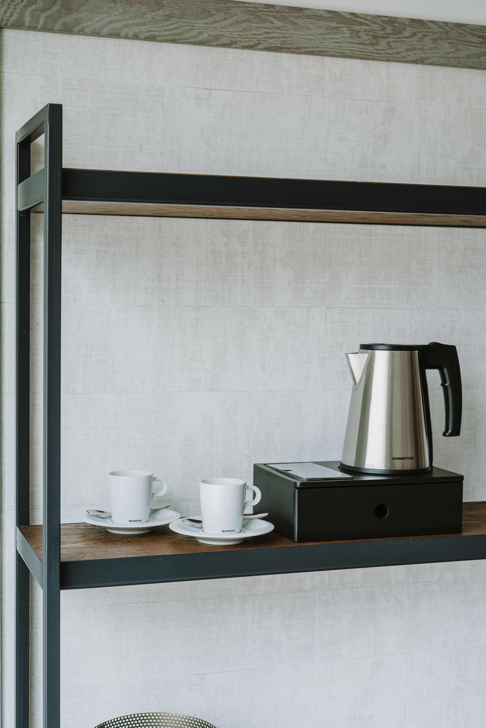 A kitchenette with a stainless steel kettle, two white cups, and a coffee maker on a wooden shelf.