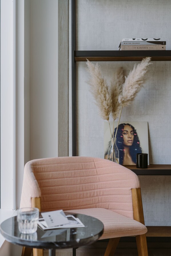 Un rincón acogedor con un sillón mullido, una mesita redonda con un vaso de agua, un florero con plumas de pampa y una estantería con libros y un cuadro enmarcado.