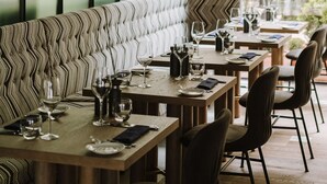 Un restaurante con una larga banqueta, varias mesas con sillas, lámparas colgantes y grandes ventanales con vistas a árboles.