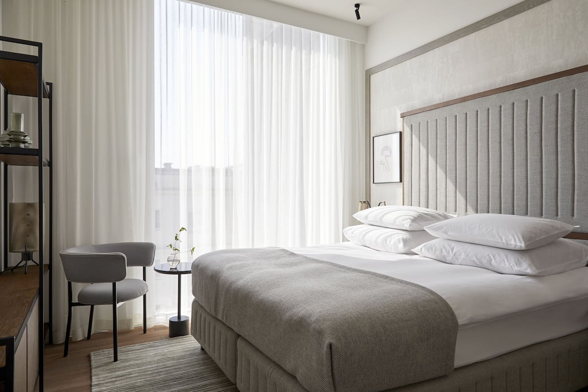 Ein Hotelzimmer mit einem großen Bett, einem Nachttisch, einem Sessel und Blick auf die Stadt durch transparente Vorhänge.