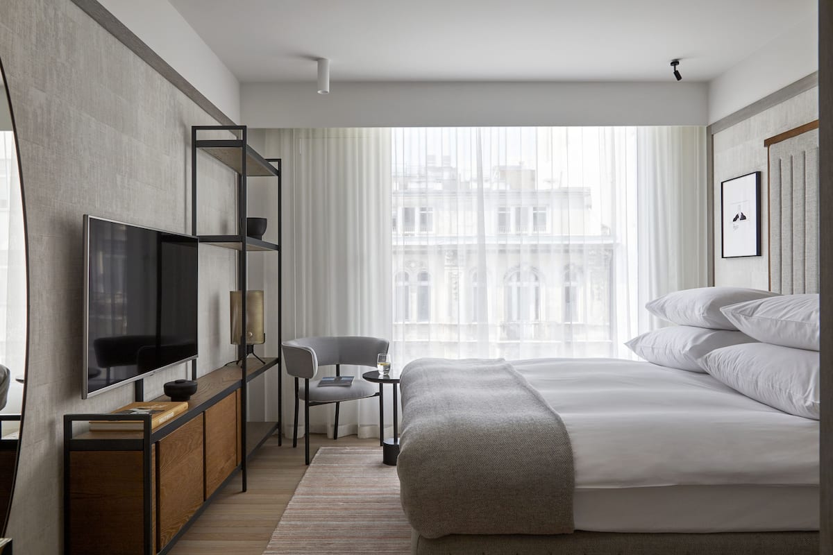 Ein modernes Hotelzimmer mit einem großen Bett, einem an einer Holzkonsole montierten Fernseher, einem kleinen Schreibtisch mit Stuhl und einem großen Fenster mit Blick auf die Stadt.