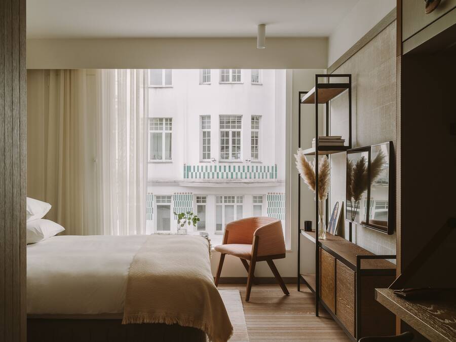 A hotel room with a bed, a chair, a wooden desk, and a view of a building with balconies.