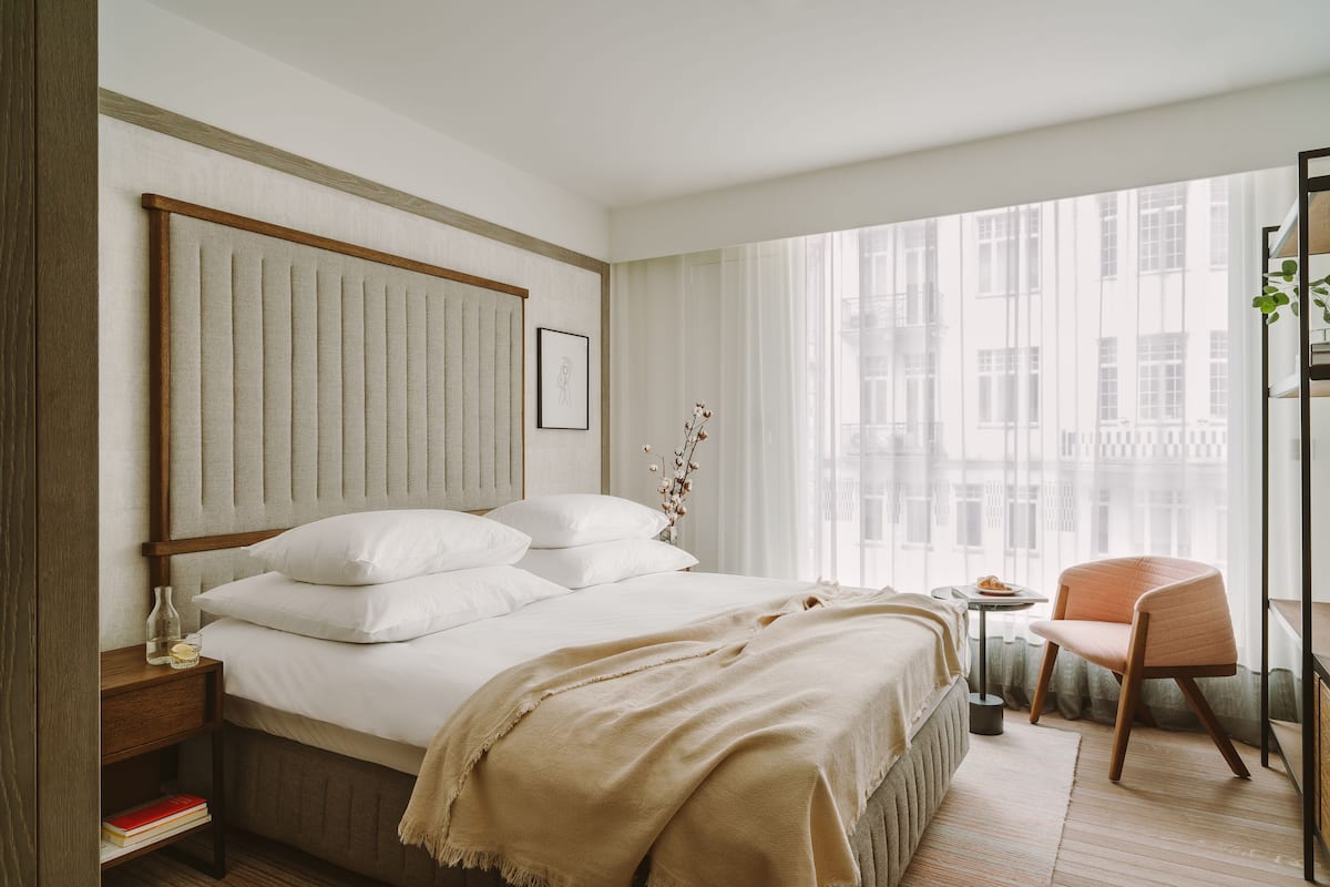 Ein modernes Hotelzimmer mit einem großen Bett, einem Nachttisch mit Lampe, einem Sessel und Blick auf die Stadt durch große Fenster.