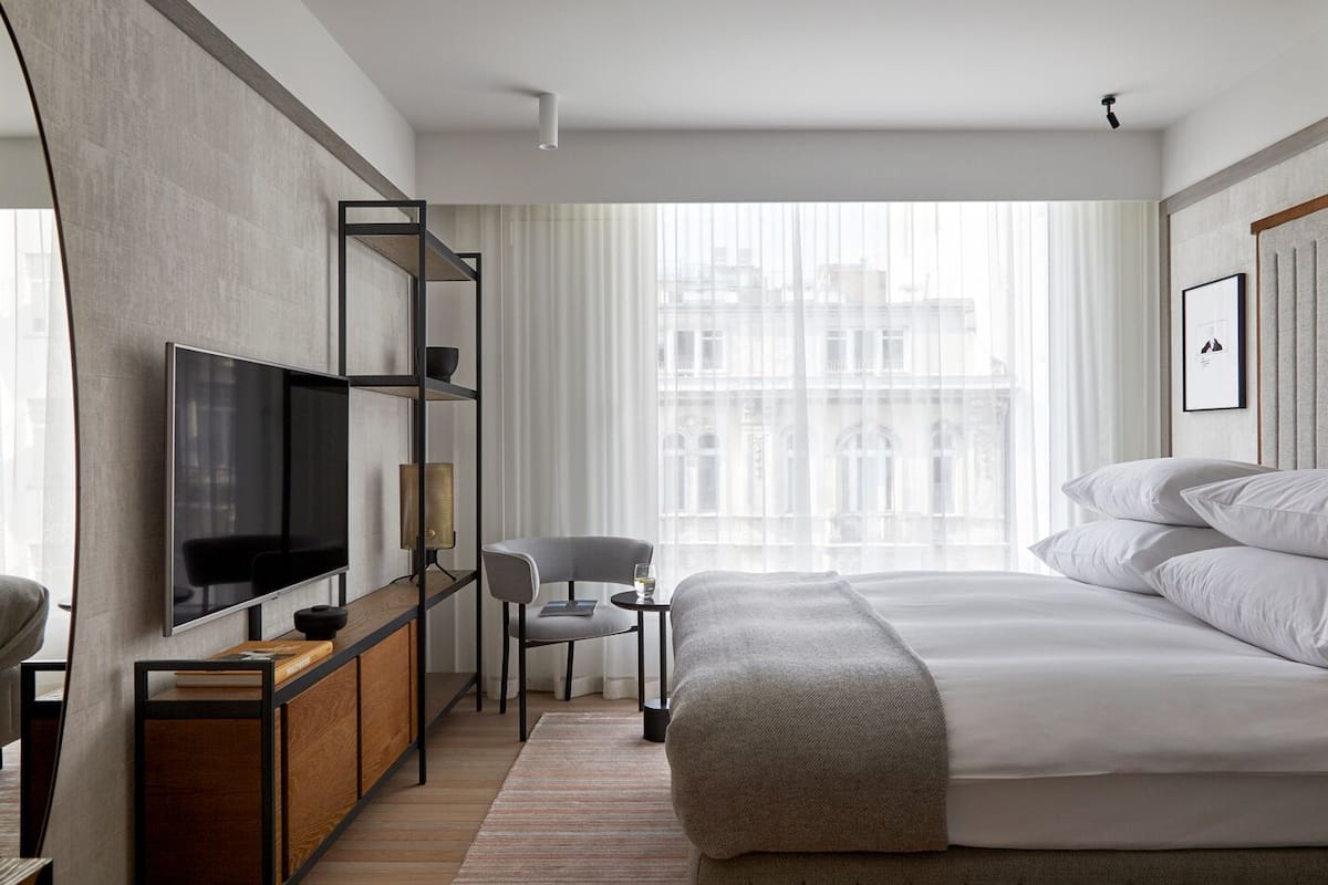 Ein modernes Hotelzimmer mit einem großen Bett, einem Fernseher, einem Schreibtisch mit Stuhl und Blick auf Gebäude durch transparente Vorhänge.