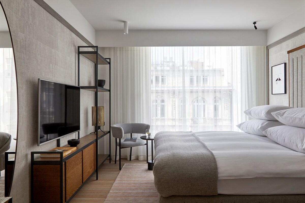 Una habitación de hotel moderna con una cama grande, un televisor, un escritorio con silla y vistas a edificios a través de cortinas translúcidas.