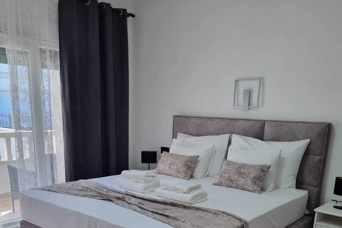 Un lit bien fait, avec une tête de lit grise, du linge de lit blanc et un plaid gris. On aperçoit une table de chevet avec une lampe et un tableau encadré au mur.