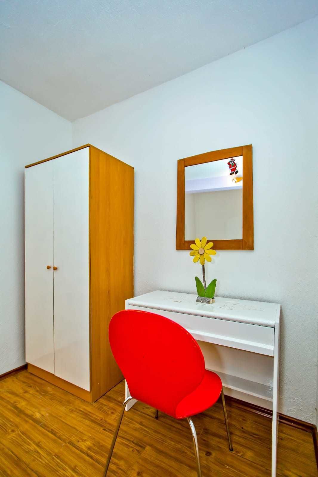 A room with a white wardrobe, a wooden door, a white desk with a mirror, a red chair, and a small yellow flower on the desk.
