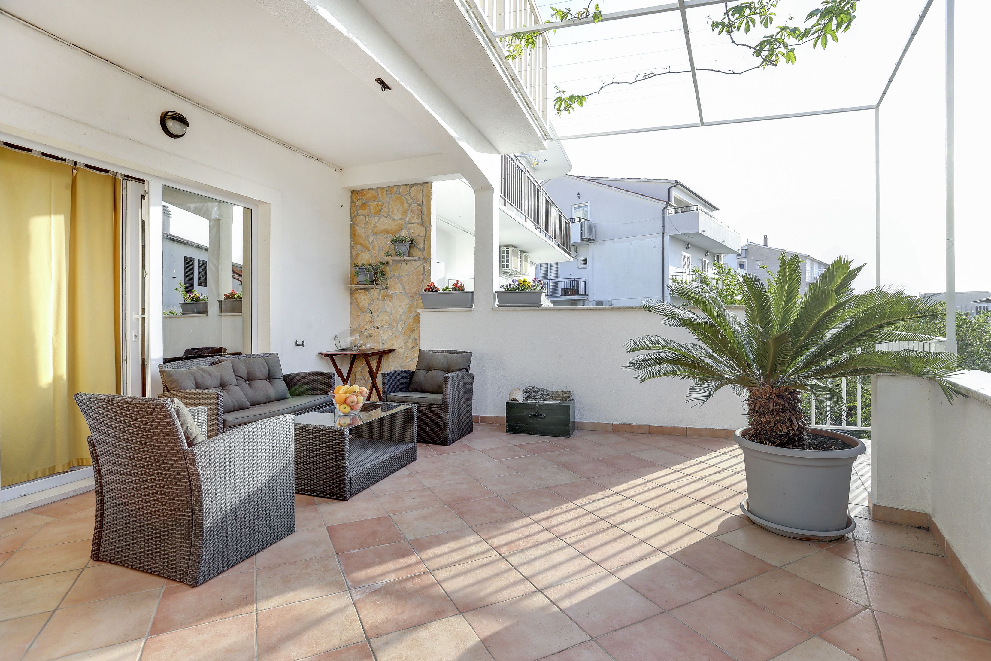 Un balcón amplio con mobiliario de mimbre, una palmera en maceta y vistas a edificios cercanos.