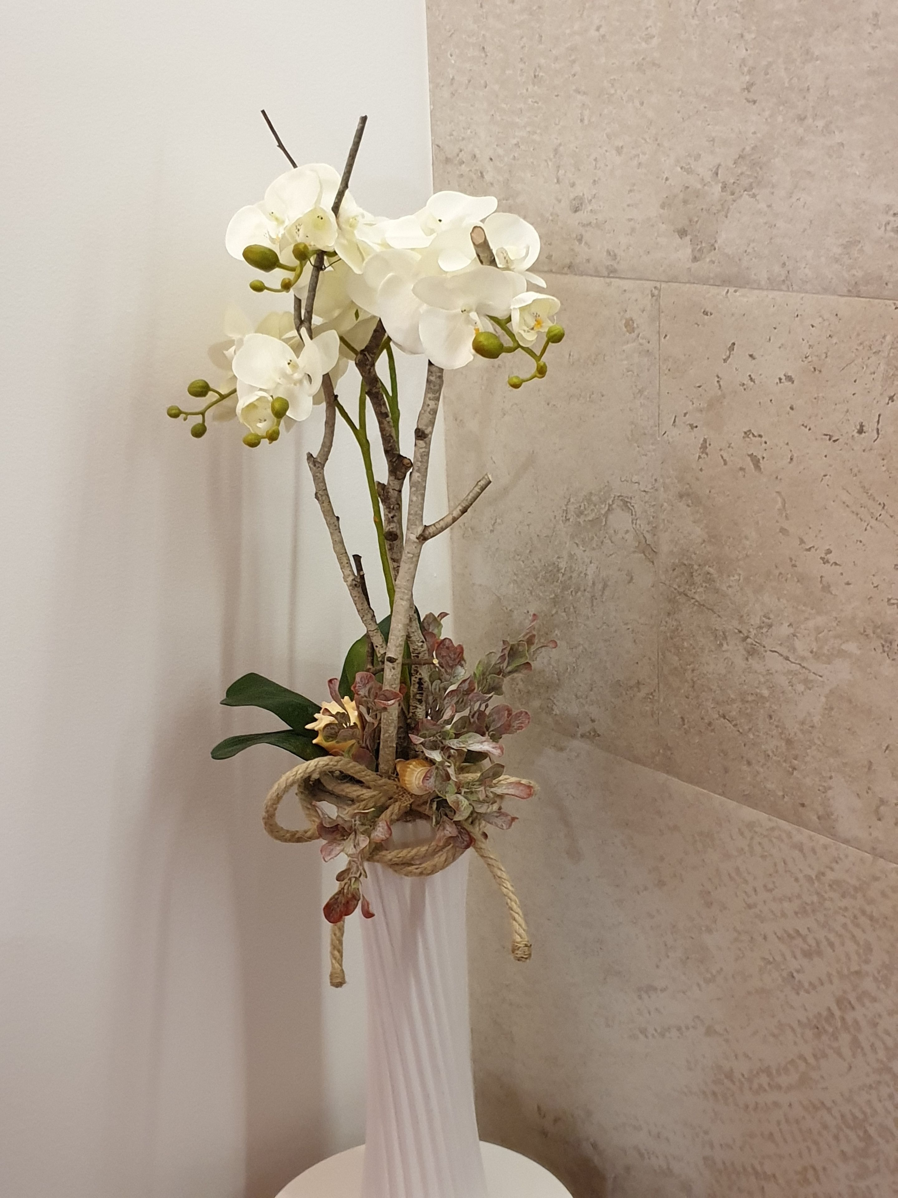 Egy fehér virágváz, melyben fehér orchideákból, zöld levelekből és dekoratív elemekből – például egy kagylóból és egy zsinórból – álló virágkompozíció található.