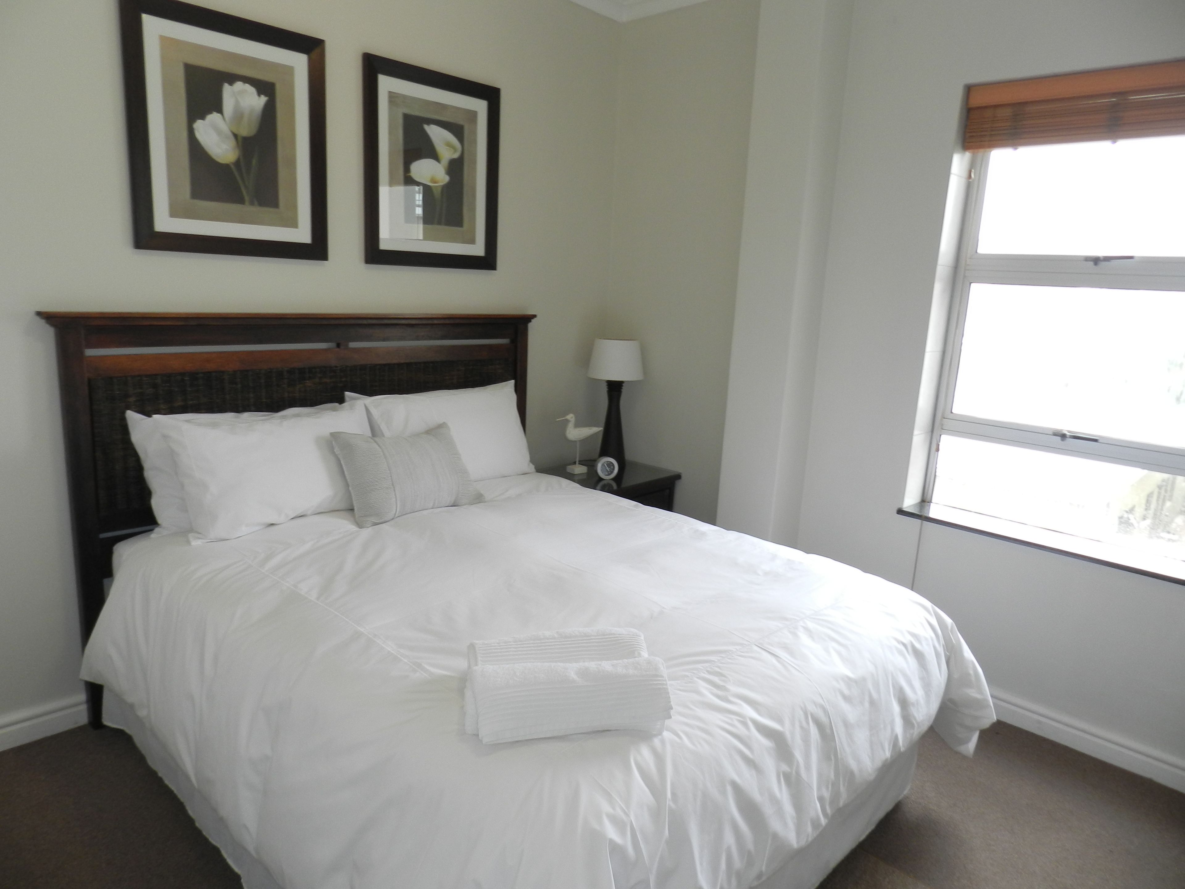 A neatly made bed with white linens and pillows, a wooden headboard, a nightstand with a lamp, and two framed floral pictures on the wall.