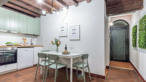 A kitchen with a dining table, chairs, and a refrigerator. There are framed pictures on the wall and a door with a glass panel.