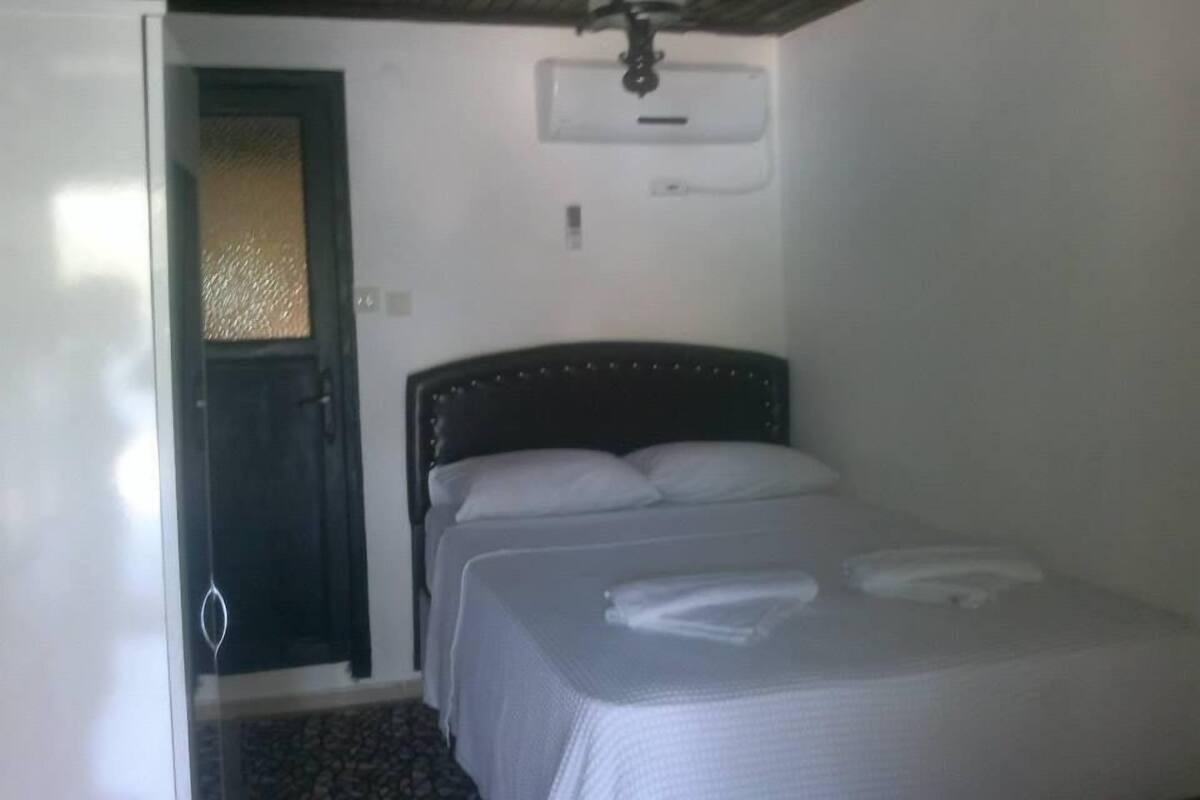 A small, single-bed room with a dark headboard, a ceiling fan, and a door leading to another room.