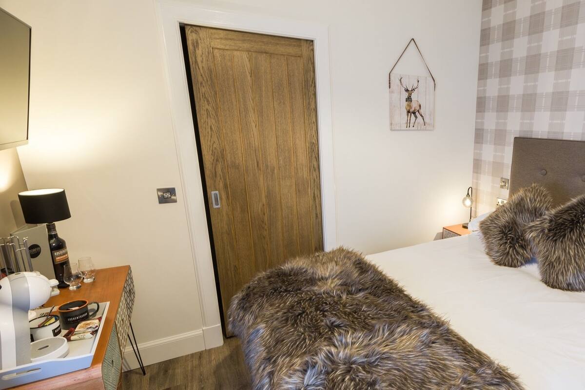 A hotel room with a wooden door, a bed with two fluffy pillows, a nightstand with a lamp, and a wall-mounted deer picture.