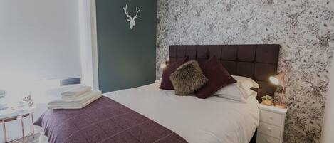 A neatly made bed with a headboard, a decorative deer wall piece, a nightstand with a lamp, and a floral wallpaper.