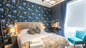 Ein ordentlich gemachtes Bett mit Kissen, einer Nachttischlampe und einem Stapel Handtücher. Rechts vom Bett befindet sich ein Stuhl und ein Spiegel.