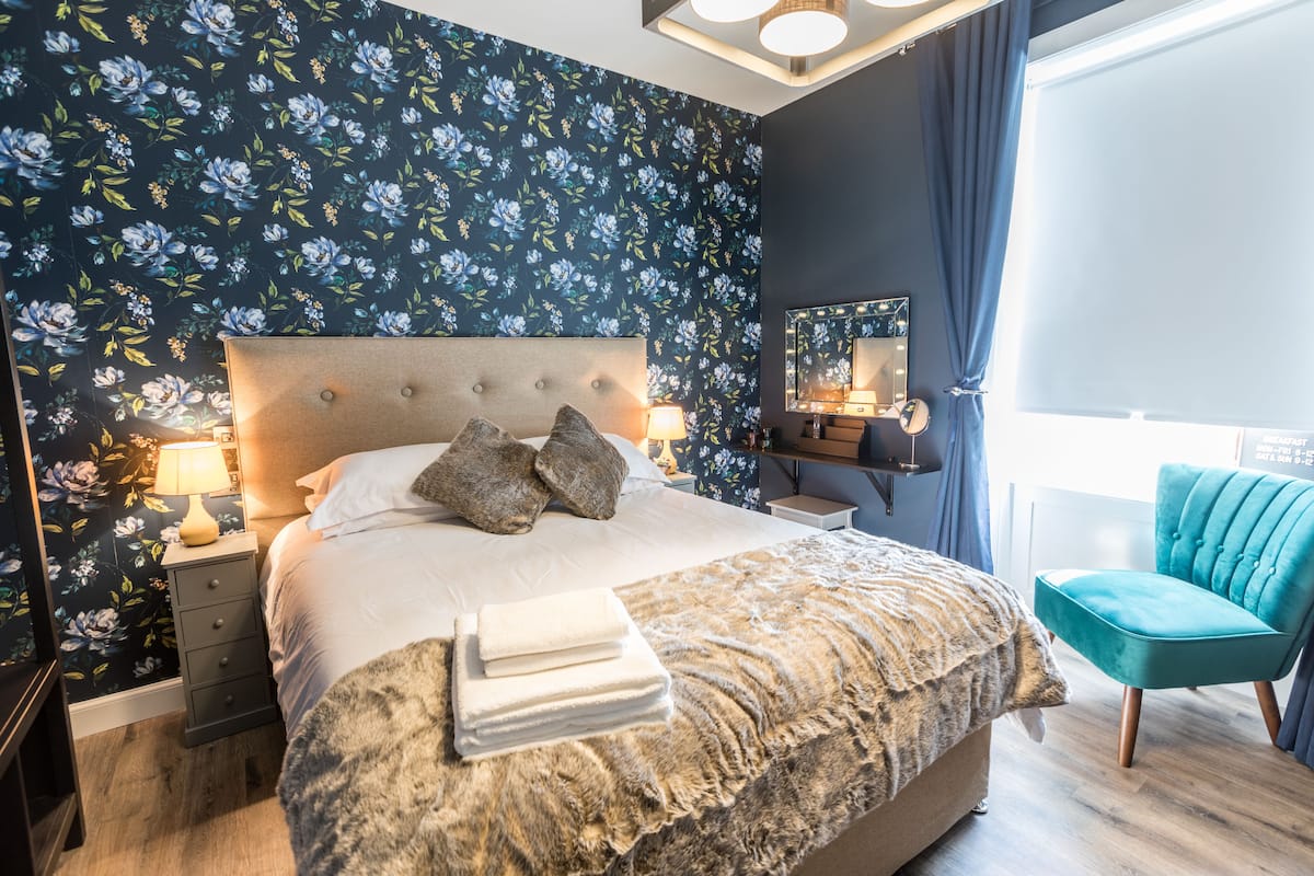 A neatly made bed with pillows, a bedside lamp, and a stack of towels. There is a chair and a mirror on the right side of the bed.