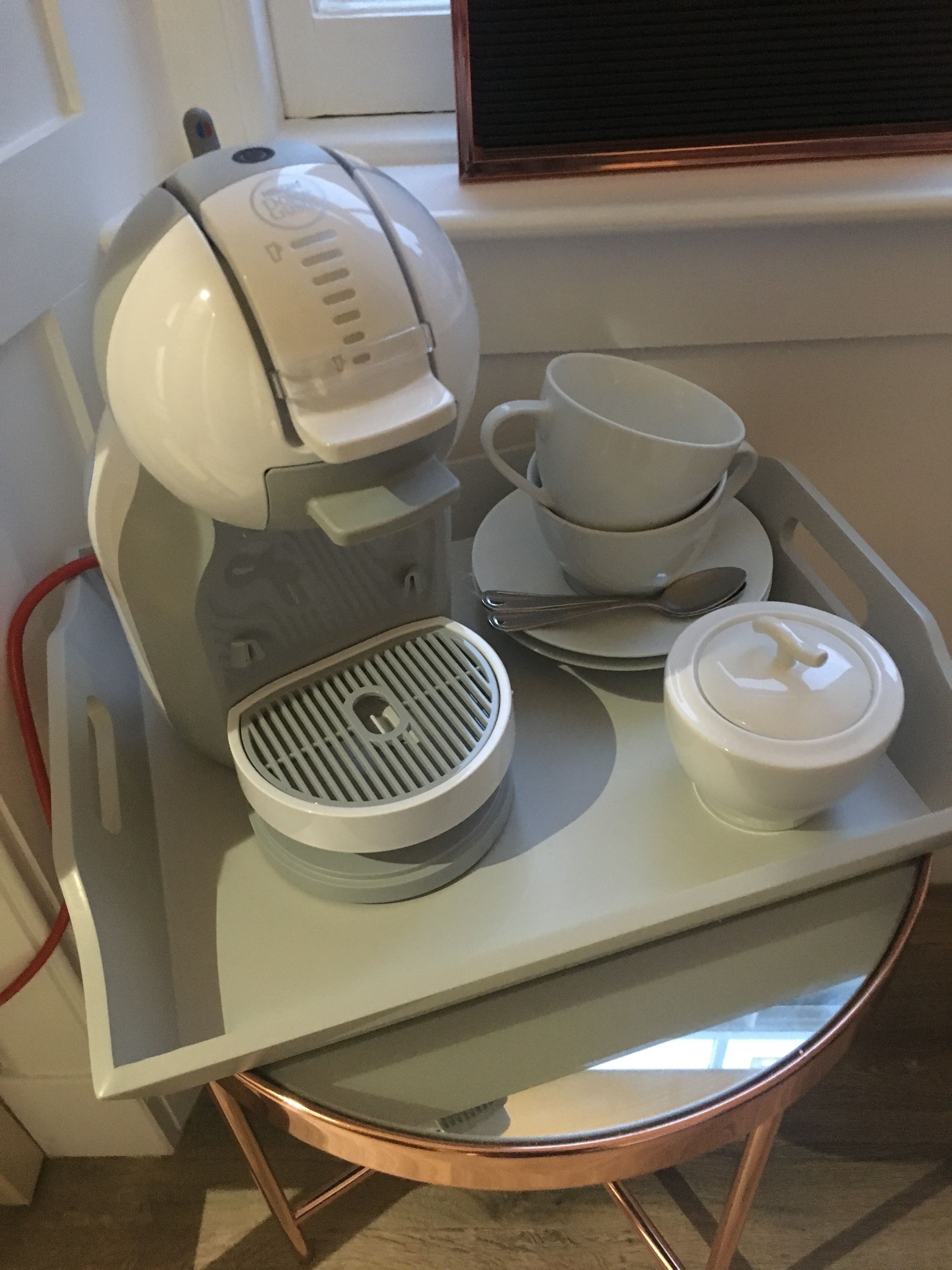 Une machine à café, des tasses et un plateau sont posés sur une table près d’une fenêtre.