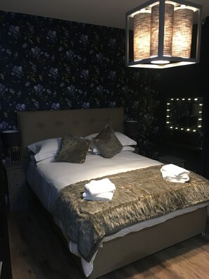 A bedroom with a large bed, a headboard, a bedside lamp, a mirror with lights, and a floral wallpaper.