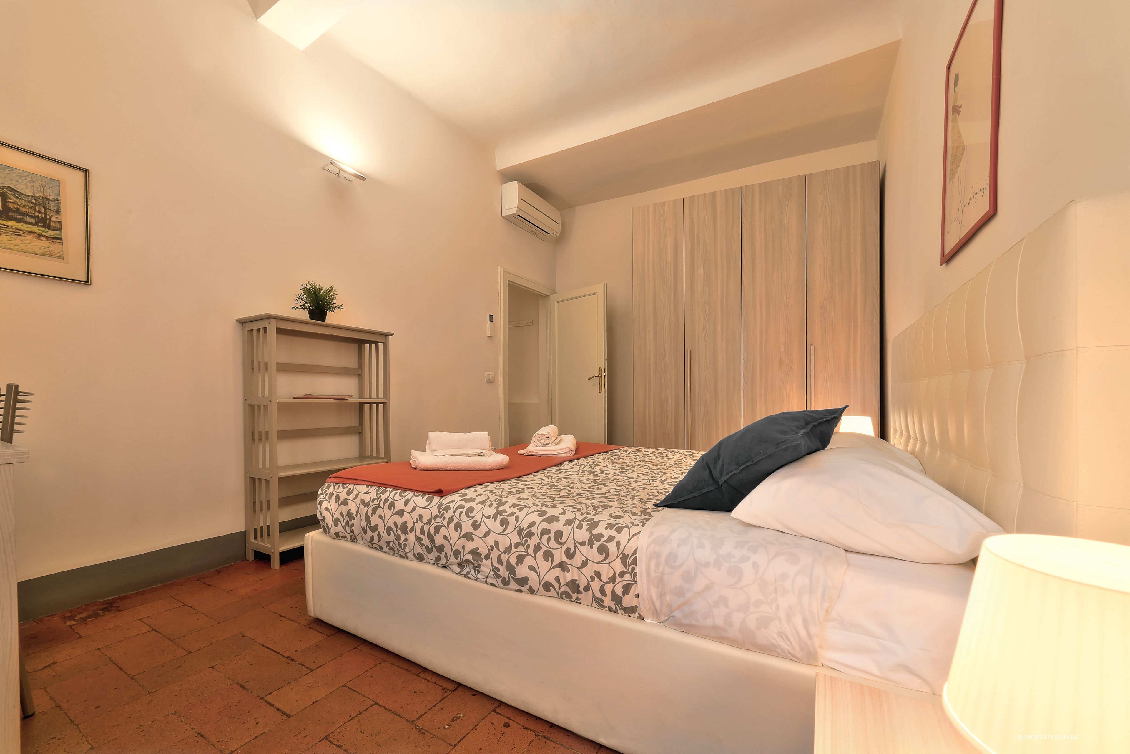 A bedroom with a bed, a bedside table, a wardrobe, a shelf unit, and a wall-mounted air conditioner.