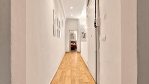 Um corredor estreito com soalho de madeira e paredes brancas, que conduz a um quarto com uma cama e uma porta.