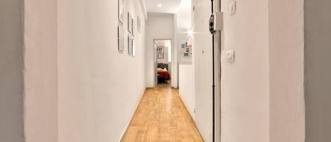 Un pasillo estrecho con suelo de madera y paredes blancas, que conduce a una habitación con una cama y una puerta.