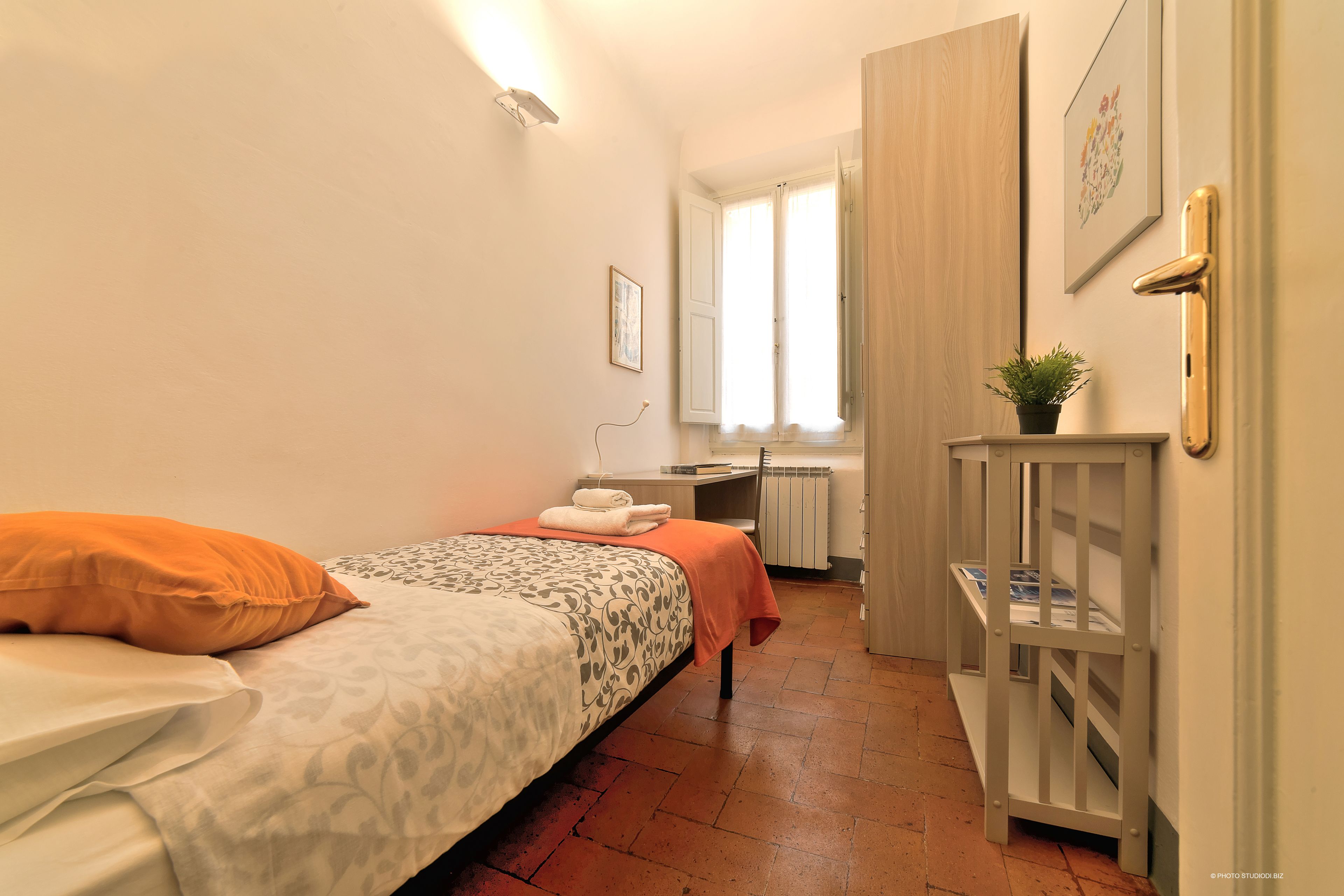 A bedroom with a bed, a desk, a radiator, and a small plant.