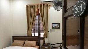 A bedroom with a bed, a nightstand, a chair, a window with curtains, a wall-mounted fan, a thermostat, and a door with a number plate. - MaiLy Hostel (Hue)