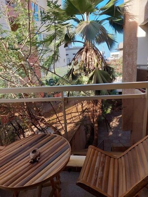 Un balcón con dos mesas de madera, una con un objeto pequeño encima, y una silla. Se observa una palmera y otra vegetación al fondo.