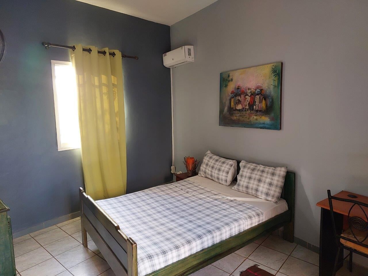 A bedroom with a bed, a window with curtains, a painting on the wall, and a small table.