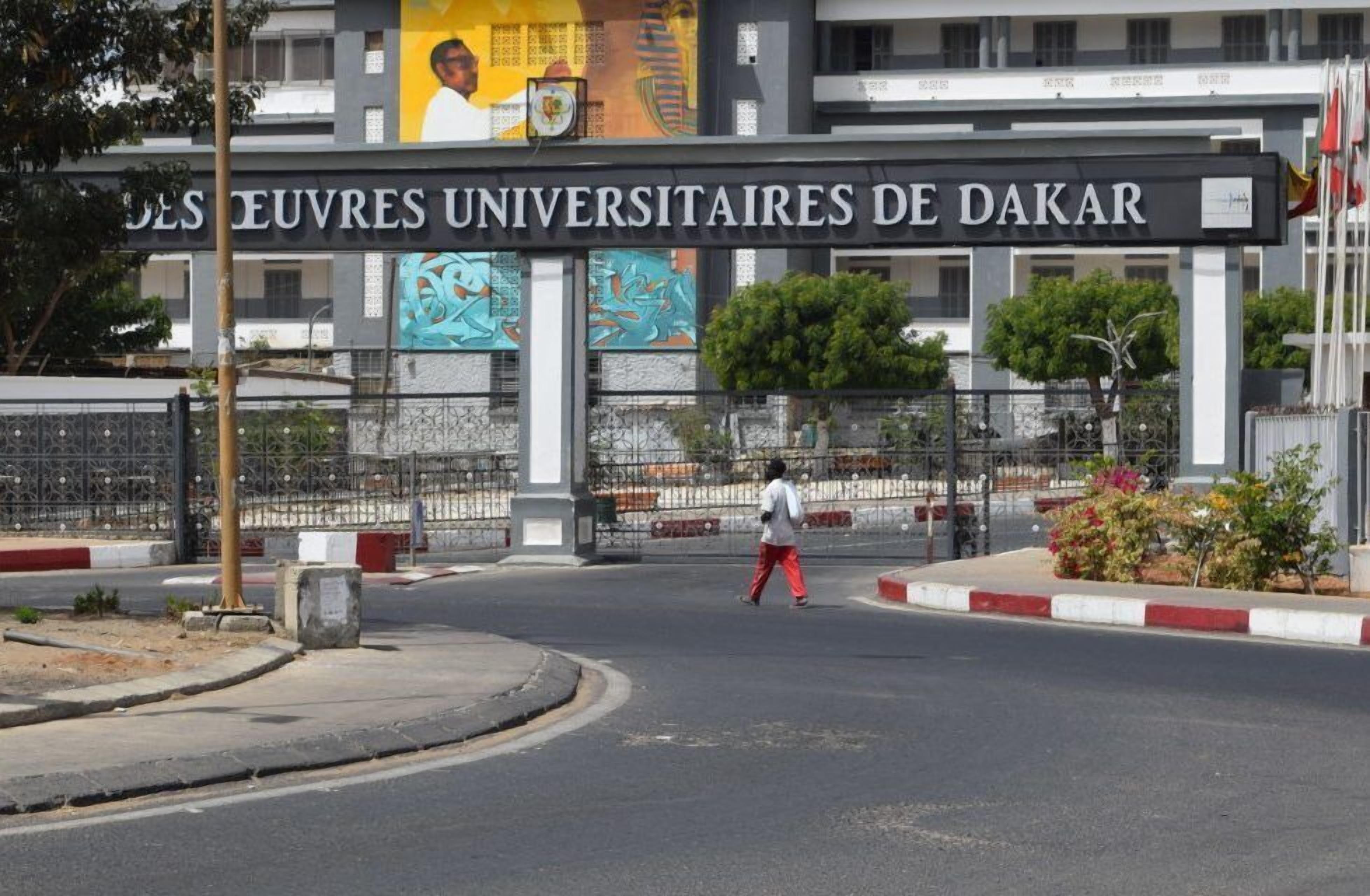 The entrance to the Universitaires De Dakar campus with a large sign, a pedestrian crossing, and a security gate.