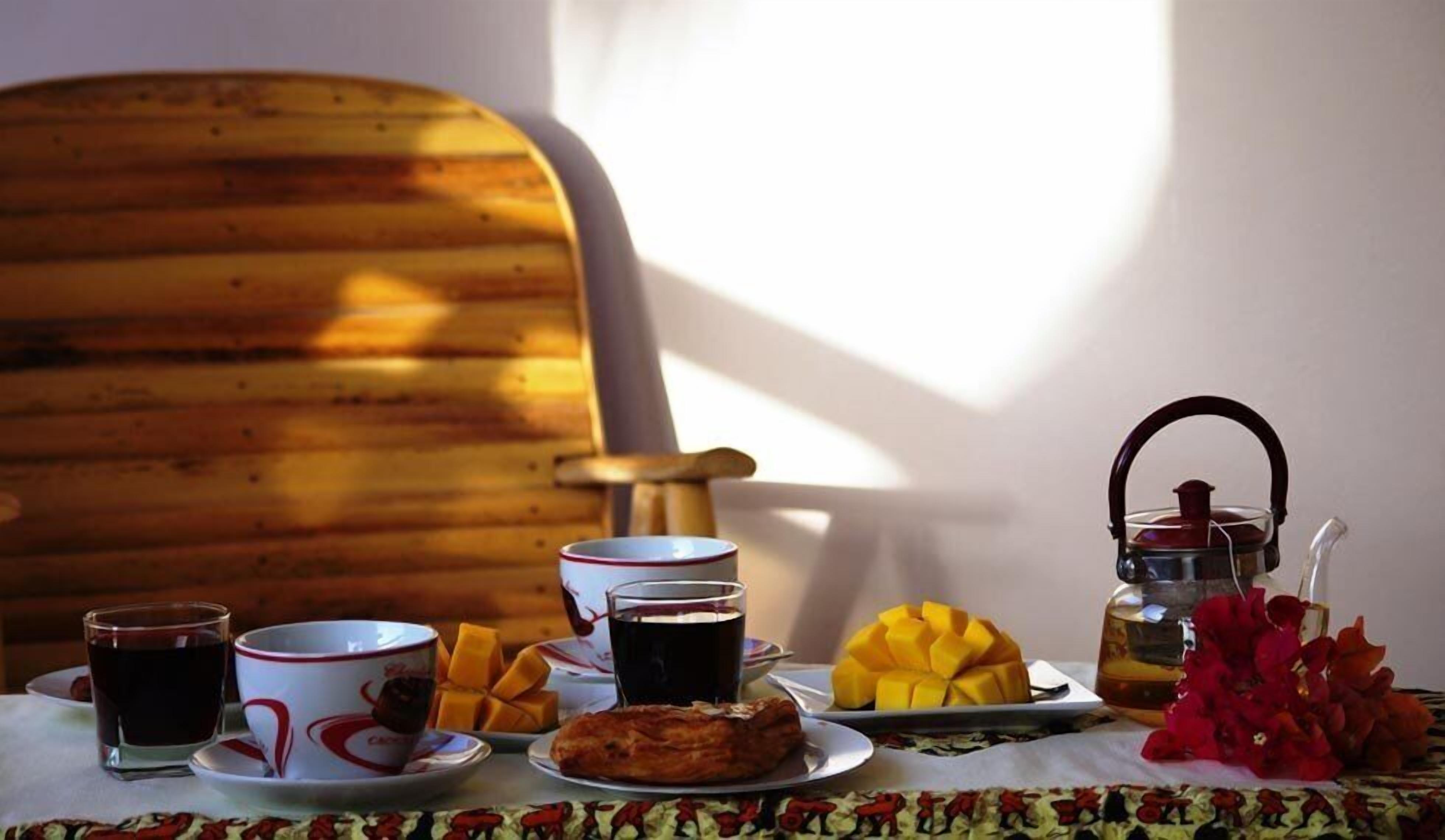 A table with a plate of food, a glass of dark liquid, a cup with a red rim, and a teapot.