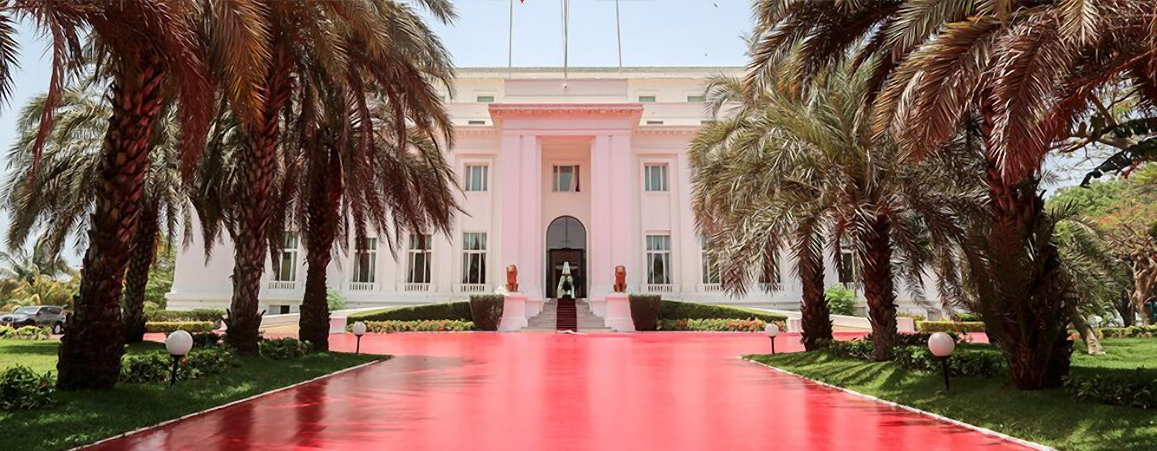 Un edificio imponente con una entrada central, flanqueado por palmeras y un sendero rojo que conduce a la entrada.