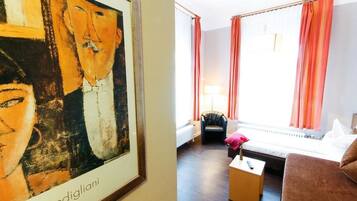 Ein Zimmer mit einem Bett, einer Couch, einem kleinen Tisch und einem Stuhl. An der Wand hängt ein Bild und es gibt ein Fenster mit Vorhängen.