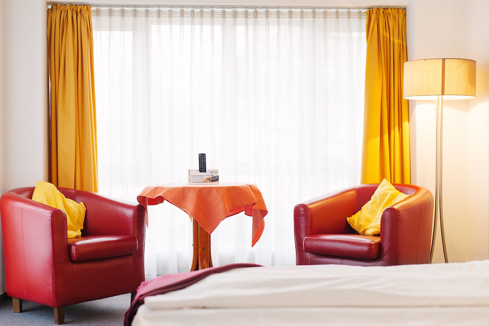 Habitación de hotel con dos sillones de cuero rojo, una mesita con mantel, una lámpara y una cama.