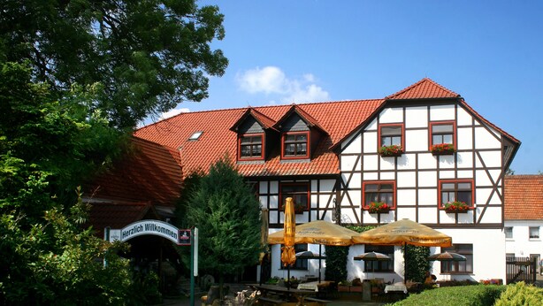 A traditional half-timbered building with a red-tiled roof, outdoor seating, and a sign reading "Herzlich Willkommen". - Pension Weidemühle (Kelbra)