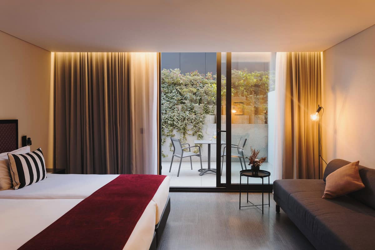 A hotel room with a bed, a sofa, a small table with chairs, and a view of an outdoor area with plants and a table.