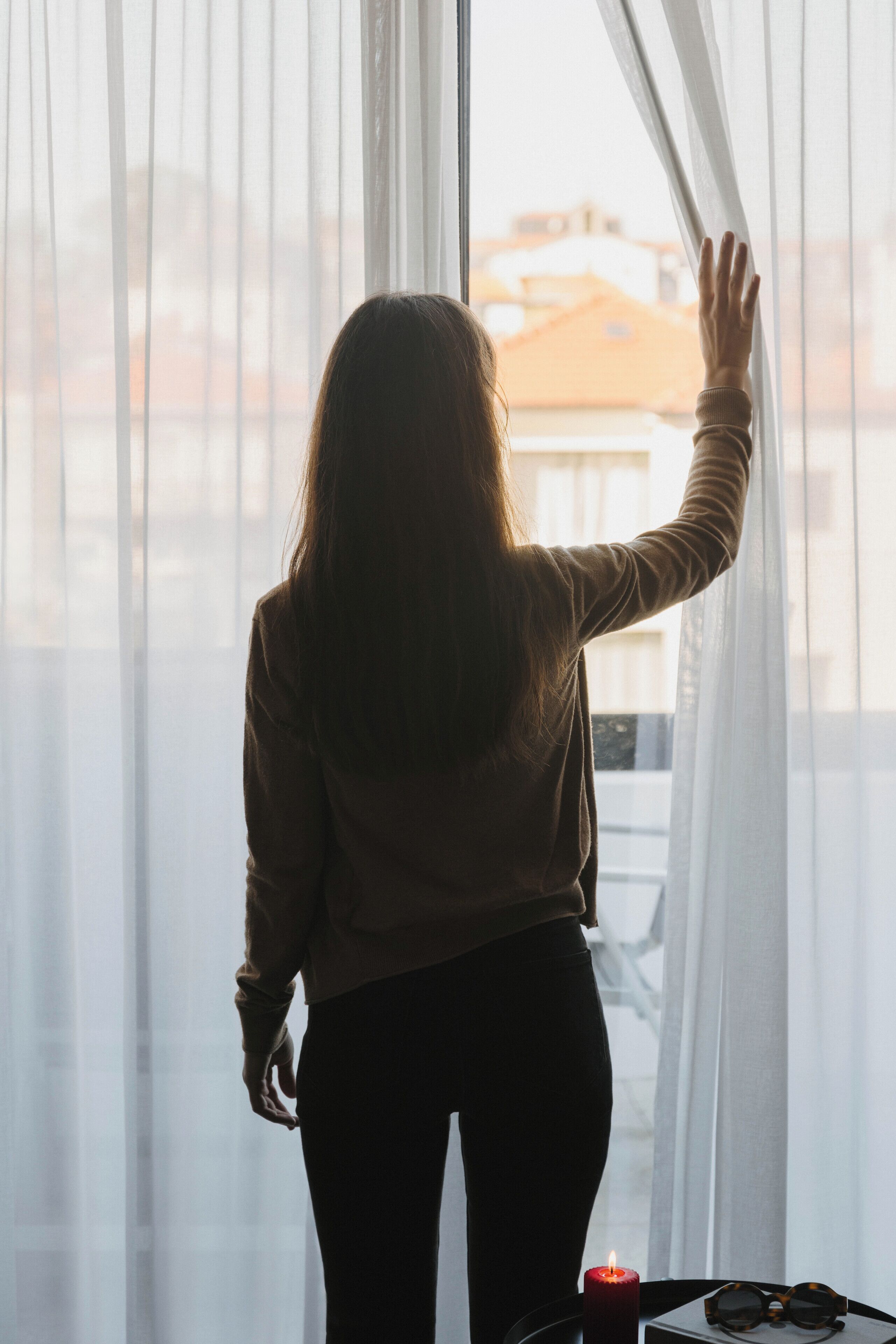 A person standing by a window with sheer curtains, a candle, and sunglasses on a table.