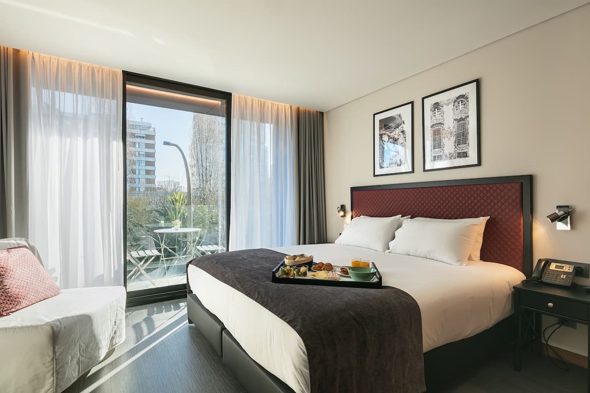 A hotel room with a large bed, a small table with breakfast, a chair, a bedside table with a phone, and a window with curtains.