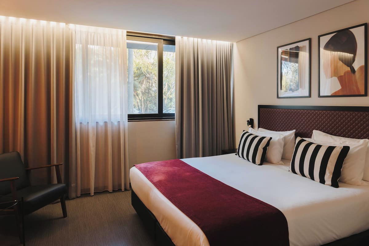 A hotel room with a large bed, a chair, a window with curtains, and two framed pictures on the wall.