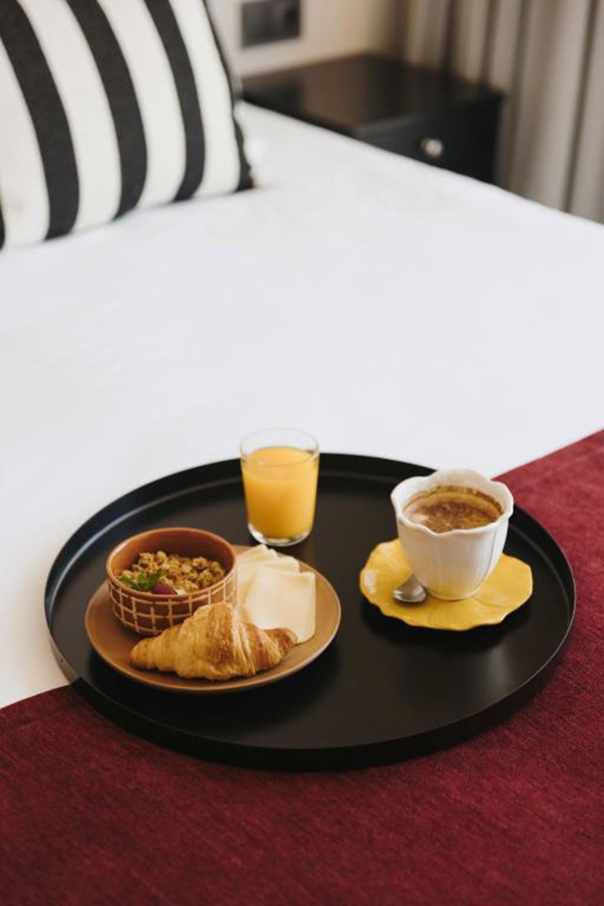 A breakfast tray with croissants, a bowl of cereal, a glass of orange juice, and a cup of coffee on a bed with a striped pillow.