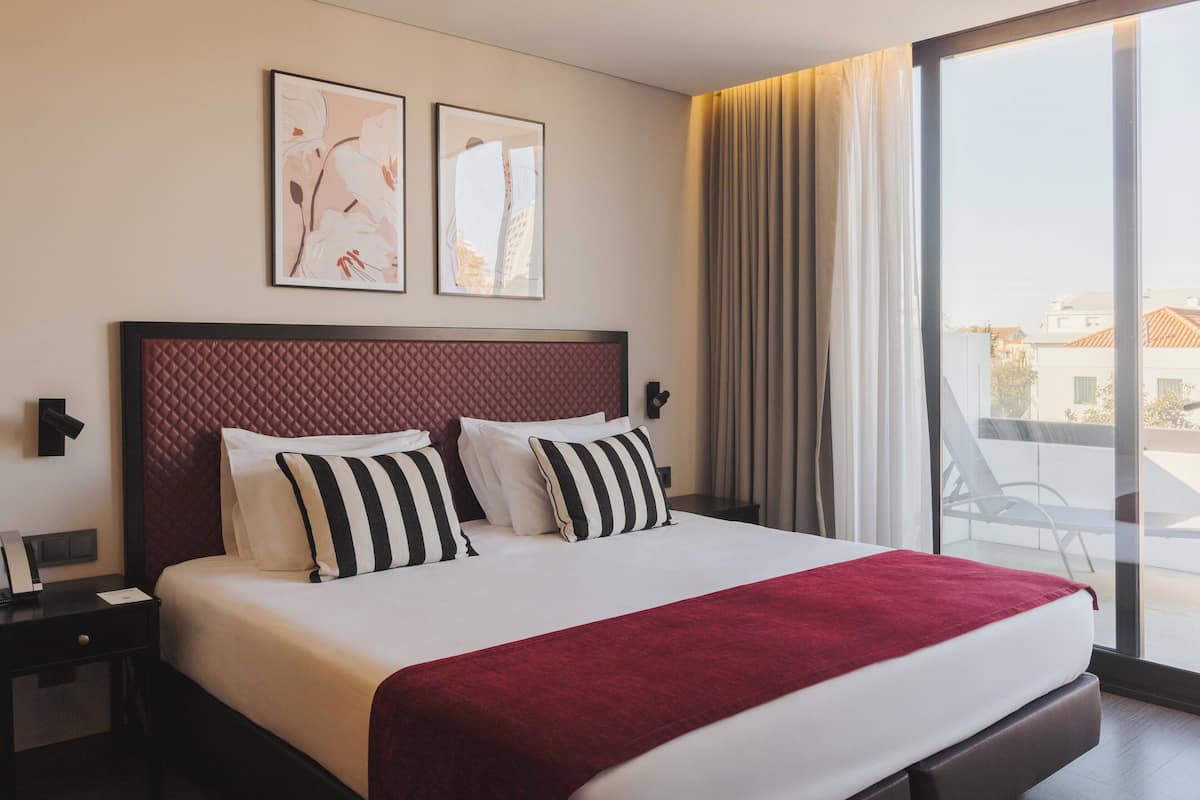 A hotel room with a large bed, a red headboard, two framed artworks on the wall, and a view of the outside through a large window.