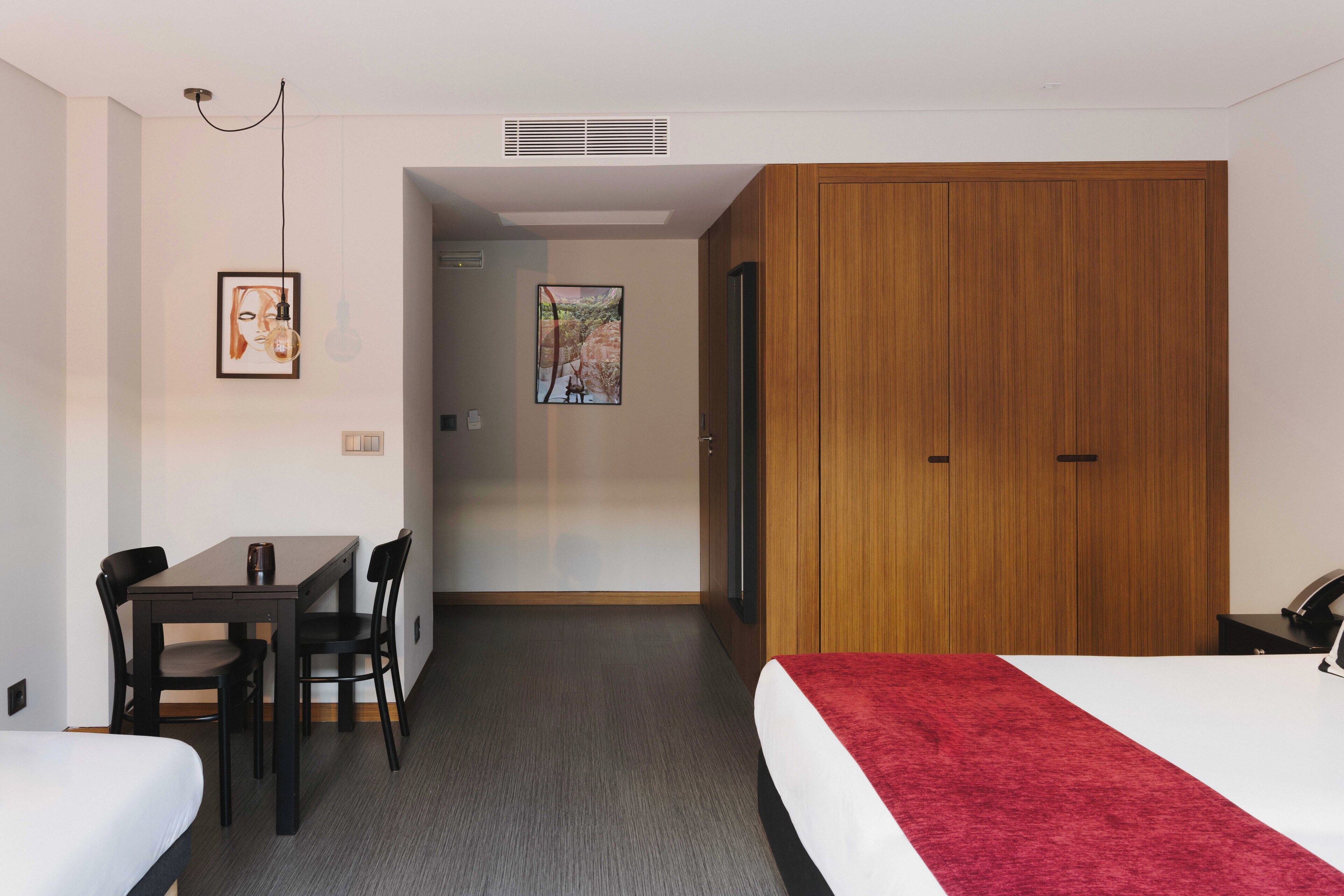 Hotellihuone, jossa on sänky, työpöytä tuoleineen, puinen vaatekaappi ja kaksi kehystettyä kuvaa seinällä.