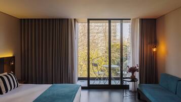 A hotel room with a bed, a sofa, a small table with a vase, and a balcony with chairs and a table.